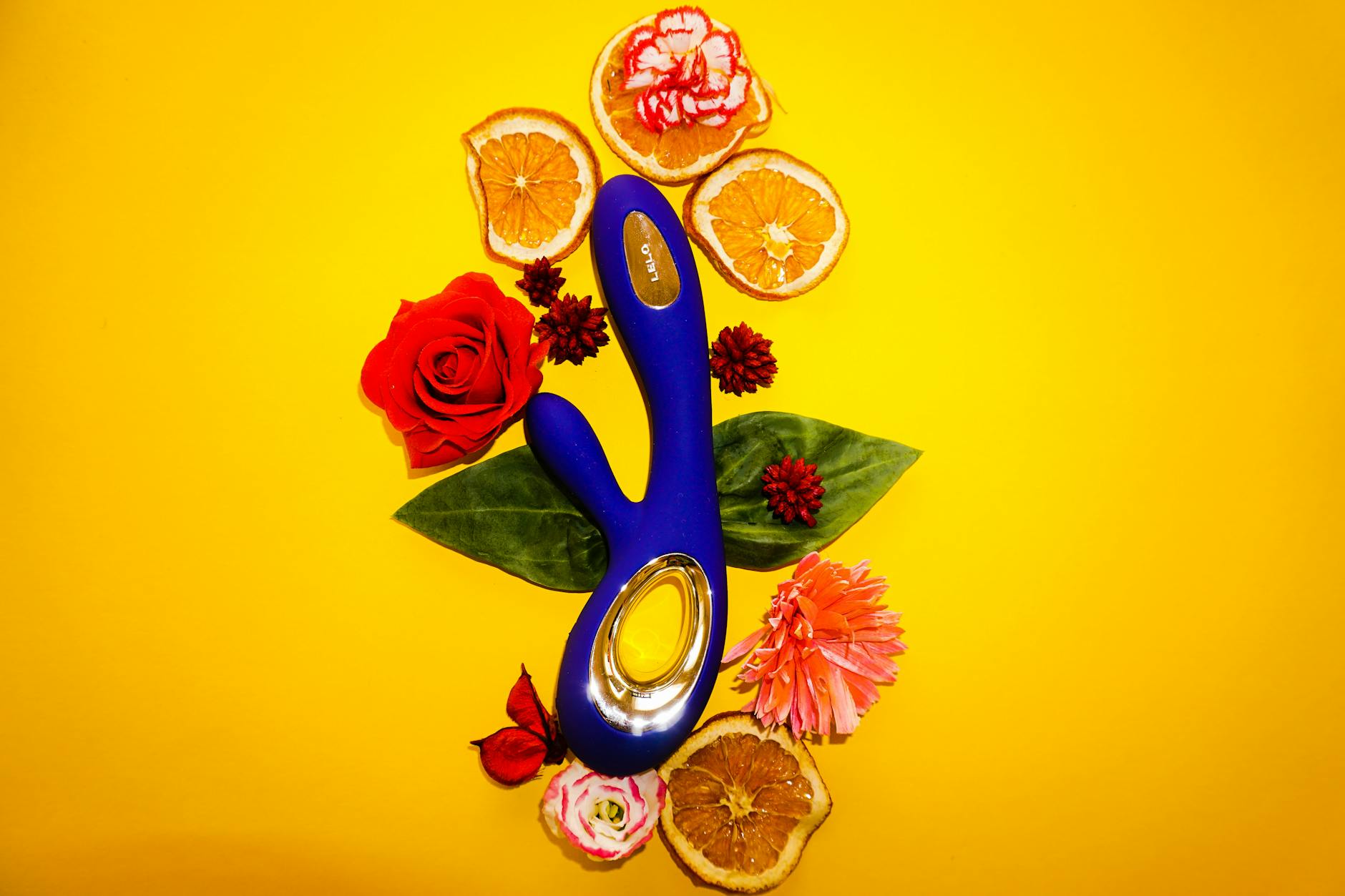 Top view of a blue vibrator surrounded by flowers and orange slices on a yellow background