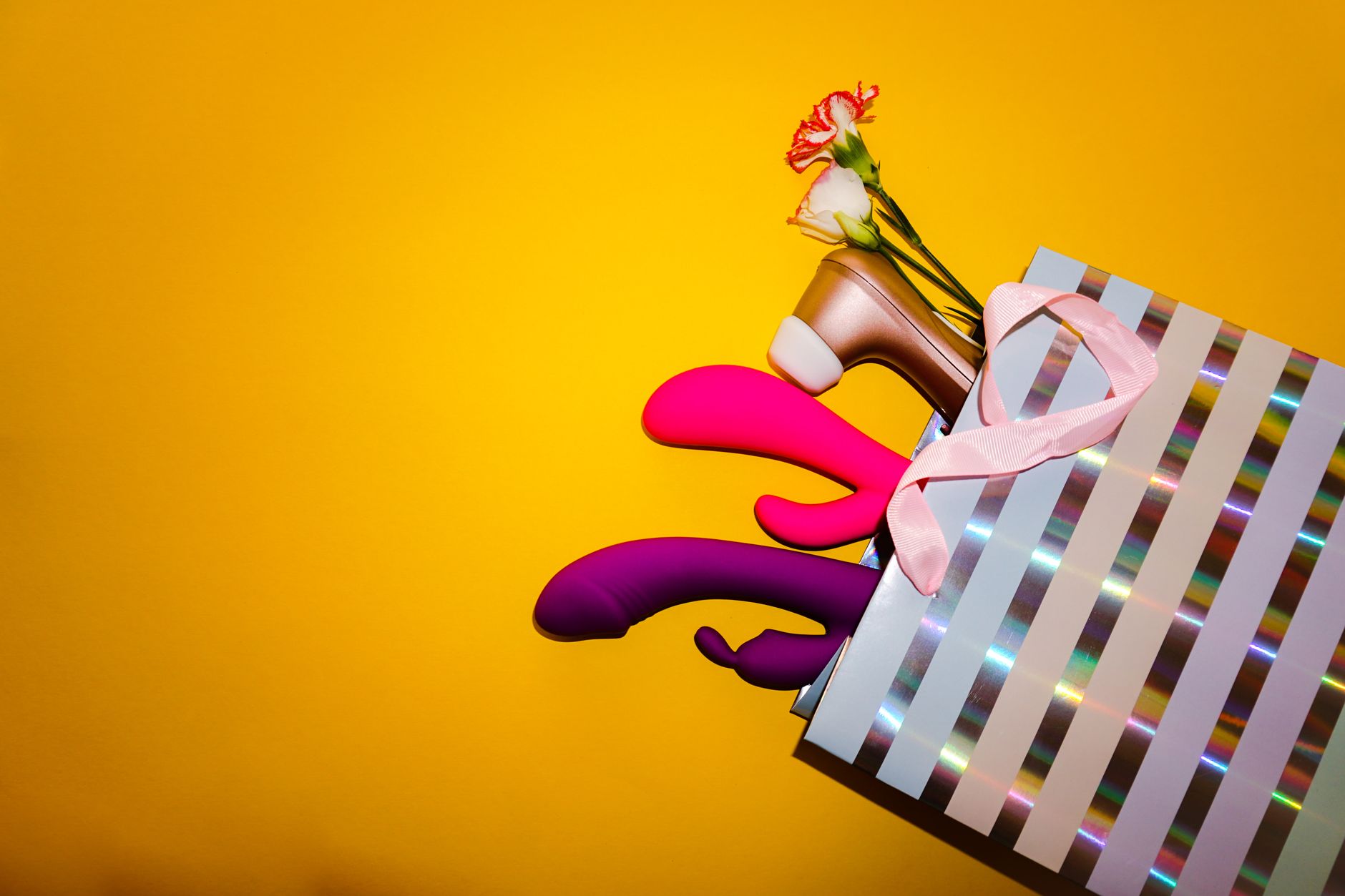 Colorful vibrators with flowers in a holographic gift bag, set against a bold yellow background